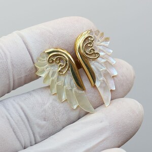 May include: A pair of gold-colored wing-shaped earrings with iridescent, pearly white feathers. The earrings have a decorative swirl design at the top and are set against a light background.