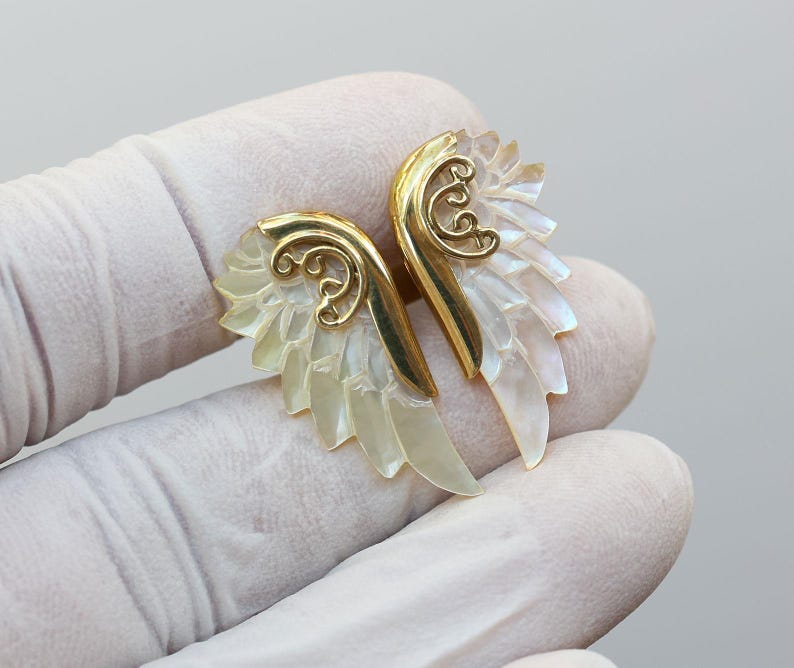 May include: A pair of gold-colored wing-shaped earrings with iridescent, pearly white feathers. The earrings have a decorative swirl design at the top and are set against a light background.
