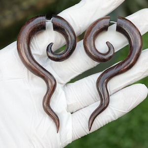 May include: A pair of dark brown wooden spiral earrings. The earrings have a curved, elongated design with a smooth, polished finish. The spiral shape tapers to a point at the bottom. The earrings are held together with a silver-coloured post.