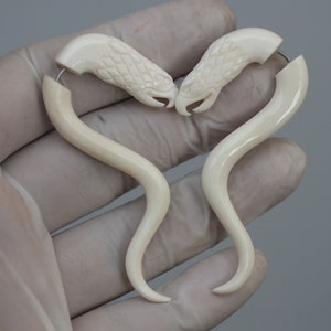 May include: A pair of white bone earrings shaped like a snake with a carved head. The earrings are curved and have a hook closure.