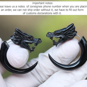 Op de afbeelding: Een paar zwarte drakenkop-oorbellen. De oorbellen zijn gemaakt van hoorn en hebben een gebogen vorm. De drakenkoppen zijn gedetailleerd gesneden en hebben scherpe tanden.