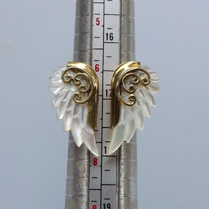 May include: A pair of gold-toned wing-shaped earrings with iridescent white mother-of-pearl feathers. The earrings have a decorative swirl design and are displayed on a silver measuring tool.
