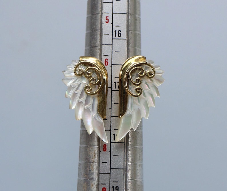 May include: A pair of gold-toned wing-shaped earrings with iridescent white mother-of-pearl feathers. The earrings have a decorative swirl design and are displayed on a silver measuring tool.