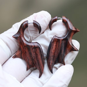 Op de afbeelding: Een paar houten oorbellen met een donkerbruin en zwart houtnerfpatroon. De oorbellen zijn gevormd als vleugels met een zilveren metalen ring.