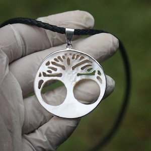 May include: A silver pendant necklace with a white mother of pearl tree of life design. The pendant is round and has a hammered texture. The necklace is on a black leather cord.