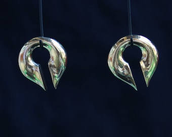May include: Two silver metal ear gauges hanging from black strings. The gauges are shaped like a teardrop with a pointed tip and a circular opening.