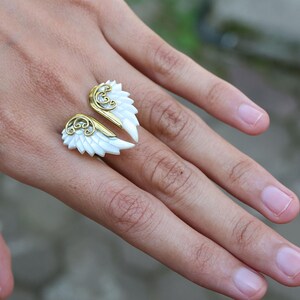 May include: A gold-toned and white wing-shaped ring. The ring features two open wings with white feathers and gold filigree accents. The ring is worn on a finger, showcasing the intricate design and craftsmanship of the jewelry.