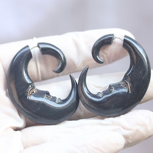 Op de afbeelding: Een paar donkergrijze halve maan oorbellen. Elke oorbel heeft een uitgesneden gezicht op het maanoppervlak. De oorbellen worden bij elkaar gehouden met een zilverkleurige metalen pin. De oorbellen zijn ontworpen om doorboorde oren te dragen.