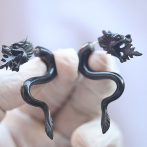 May include: A pair of black dragon head ear gauges. The gauges are made of wood and have a curved shape. The dragon heads are detailed and have sharp teeth.