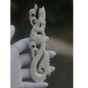 May include: A white bone carving of a traditional Maori hei tiki pendant, featuring intricate details and black eyes. The pendant is held in a gloved hand.