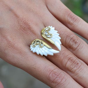 May include: A ring featuring a pair of white, feathered wings with gold-colored accents. The wings are open, creating a unique design. The ring is set on a hand, showcasing the jewelry's details and craftsmanship.