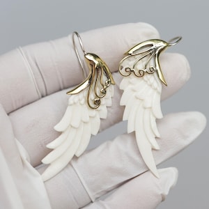 Puede incluir: Un par de pendientes de ala de ángel de hueso blanco con detalles en filigrana dorada. Los pendientes son colgantes y tienen un diseño delicado e intrincado.