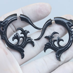 Op de afbeelding: Een paar zwarte, sierlijke oorbellen met een wervelend, bloemmotief. De oorbellen zijn gemaakt van een donker materiaal en hebben een gebogen vorm met ingewikkelde details. De oorbellen zijn ontworpen om als paar te worden gedragen.