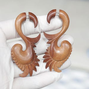 Op de afbeelding: Een paar houten oorbellen met een uniek, ingewikkeld ontwerp. De oorbellen zijn bruin en hebben een bloemmotief. Ze worden in een handschoen gehoud.