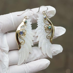 Op de afbeelding: Oorbellen met een gebeeldhouwd wit veermotief. De oorbellen hebben goudkleurige metalen accenten met wervelende filigraan en iriserende blauwgroene stenen. De oorhaken zijn zilverkleurig.