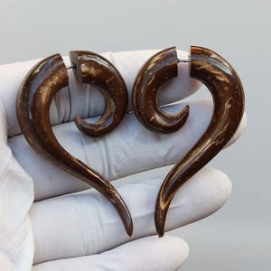 Op de afbeelding: Een paar donkerbruine, spiraalvormige oorbellen. De oorbellen zijn gemaakt van een natuurlijk materiaal, mogelijk hout of kokosnoot, en hebben een gepolijste afwerking. Het ontwerp is een gebogen, langwerpige spiraal, met een kleine pin.