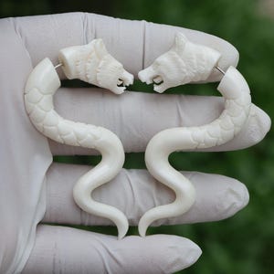 Puede incluir: Un par de pendientes de hueso blanco con forma de cabeza de lobo y cola curva. Los pendientes están hechos de hueso y tienen un color blanco natural.