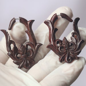 Op de afbeelding: Een paar donkerbruine houten oorbellen met ingewikkelde bloemmotieven. De oorbellen zijn hoepelvormig en hebben een zilveren metalen hoepel.