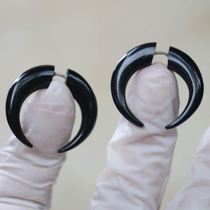 Op de afbeelding: Een paar zwarte, hoornvormige oorbellen. De oorbellen hebben een gebogen ontwerp met een spleet aan de bovenkant, bij elkaar gehouden door een zilverkleurige metalen staaf. De oorbellen zijn glanzend en lijken van een glad materiaal te zijn gemaakt.