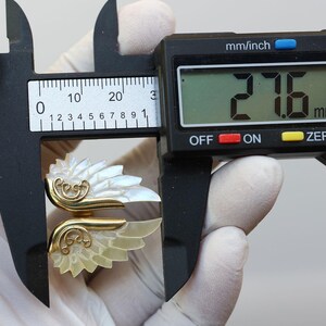 May include: A gold-toned ring with a mother-of-pearl wing design is measured with a digital caliper. The caliper displays a measurement of 27.6 mm. The ring features intricate gold filigree details.
