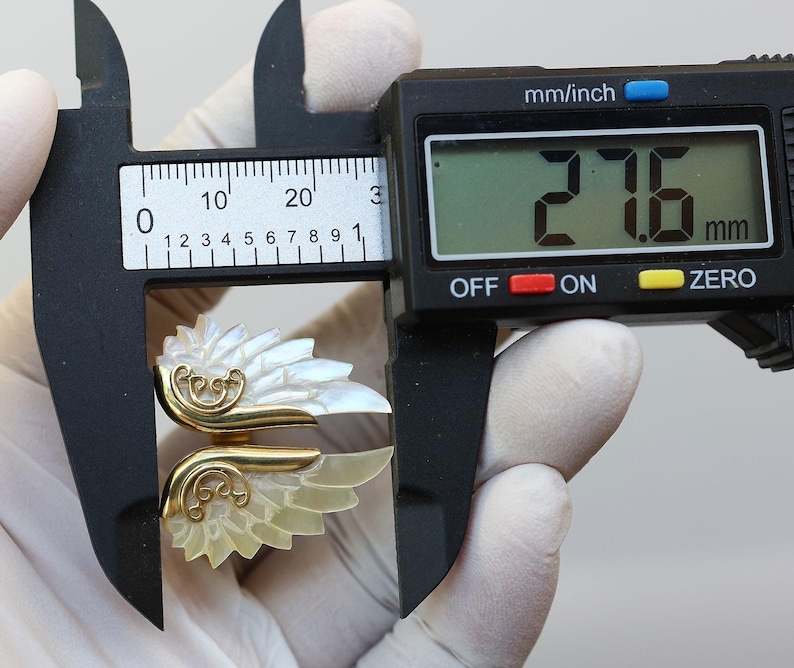 May include: A gold-toned ring with a mother-of-pearl wing design is measured with a digital caliper. The caliper displays a measurement of 27.6 mm. The ring features intricate gold filigree details.
