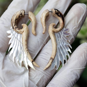 May include: A pair of handcrafted wooden earrings with a unique design. The earrings feature a light-colored wood with dark brown accents, and a wing-shaped, iridescent, mother-of-pearl element. The earrings are designed to be worn through pierced ears.