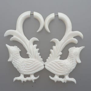 Op de afbeelding: Een paar witte oorbellen in de vorm van een haan. De oorbellen hebben een gedetailleerd ontwerp van een haan met een gebogen bovenkant en een decoratief veermotief. De oorbellen zijn gemaakt van een solide, ondoorzichtig materiaal.