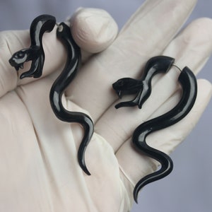 Op de afbeelding: Een paar zwarte slangvormige oorbellen. De oorbellen zijn gemaakt van glad, zwart materiaal en hebben een zilverkleurige haak.