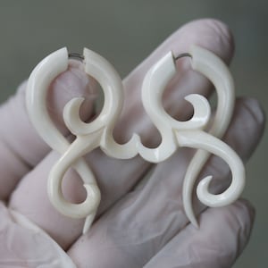 Op de afbeelding: Een paar crèmekleurige, gesneden oorbellen met een ingewikkeld, wervelend ontwerp. De oorbellen hebben een haaksluiting en zijn gemaakt van een glad, gepolijst materiaal. Het ontwerp omvat gebogen lijnen en open ruimtes.