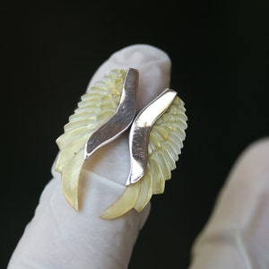 Puede incluir: Un par de pendientes de plata con un delicado diseño tallado que se asemeja a alas. Las alas están hechas de un material amarillo claro, posiblemente nácar.