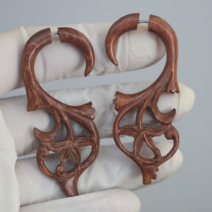 May include: A pair of wooden earrings with a floral design. The earrings are brown and have a delicate, intricate pattern. They are made of wood and have a silver metal hoop.