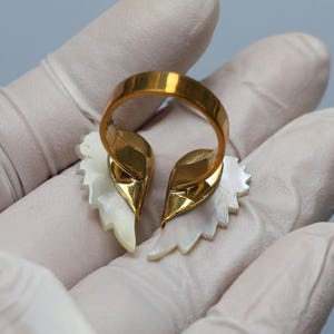 May include: A gold-toned ring featuring a split design with two stylized, white, leaf-shaped elements. The ring's band is a simple, polished gold. The leaf-shaped elements have a pearlescent sheen.