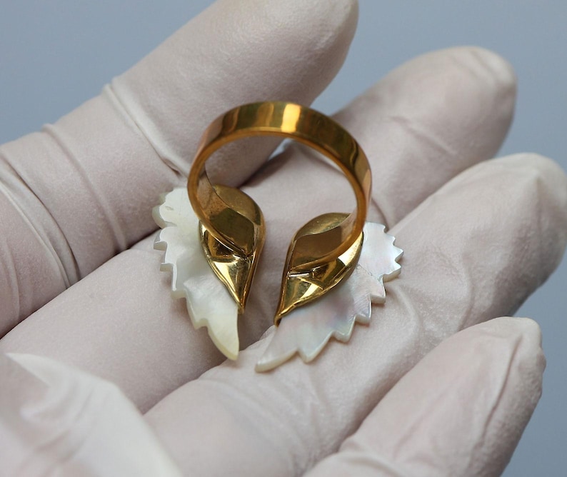 May include: A gold-toned ring featuring a split design with two stylized, white, leaf-shaped elements. The ring's band is a simple, polished gold. The leaf-shaped elements have a pearlescent sheen.