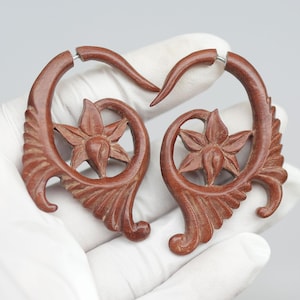 Op de afbeelding: Een paar houten oorpluggen met een bloemmotief. De pluggen zijn bruin en hebben een natuurlijke houtkleur. Ze zijn spiraalvormig met een bloem in het midden.