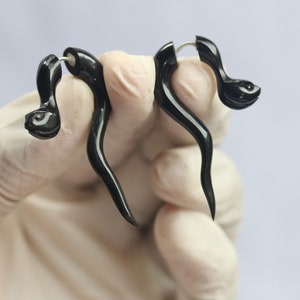 Puede incluir: Un par de pendientes negros y curvos. Los pendientes tienen un diseño en espiral con un extremo redondeado y texturizado. Los pendientes son sostenidos por una mano enguantada sobre un fondo blanco. Los pendientes están hechos de un material oscuro y brillante.