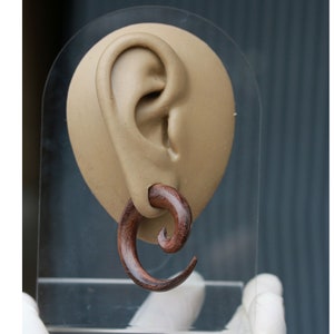 May include: A brown wooden spiral ear gauge, also known as a plug or tunnel, is shown in a display stand. The gauge is worn in a model ear.
