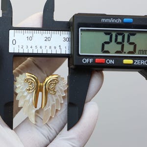 May include: A gold-colored metal and mother-of-pearl wing-shaped pendant. The pendant is measured with a digital caliper, showing a width of 29.5 mm, or approximately 1.16 inches. The wings have a detailed, layered design.