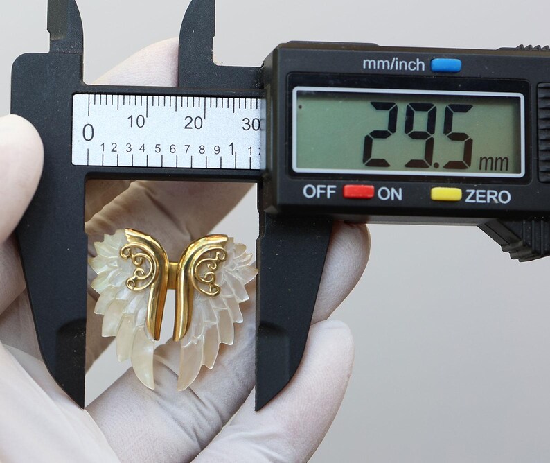 May include: A gold-colored metal and mother-of-pearl wing-shaped pendant. The pendant is measured with a digital caliper, showing a width of 29.5 mm, or approximately 1.16 inches. The wings have a detailed, layered design.
