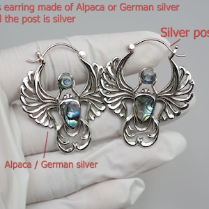 Puede incluir: Un par de pendientes plateados con un diseño de alas de ángel. Cada pendiente presenta una piedra de abulón central y el texto "this earring made of Alpaca or German silver and the post is silver". Los postes son plateados.