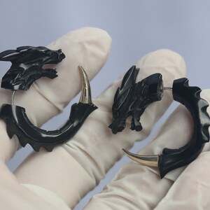 Op de afbeelding: Een paar zwarte oorbellen in de vorm van een draak. De oorbellen hebben een gedetailleerd drakenkopontwerp en een gebogen, klauwachtige vorm. De pinnen en accenten zijn zilverkleurig. De oorbellen worden gepresenteerd tegen een lichtblauwe achtergrond.