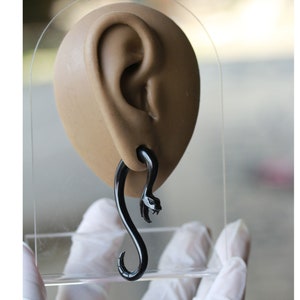 May include: Black snake shaped ear cuff. The cuff is made of a smooth, shiny material and is shaped like a snake with a curled tail. The snake's head is facing forward and has a slightly open mouth.