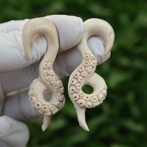 Può includere: Un paio di dilatatori per orecchie in legno bianco a forma di polpo. I dilatatori sono intagliati con dettagli intricati e hanno una finitura liscia e lucida.