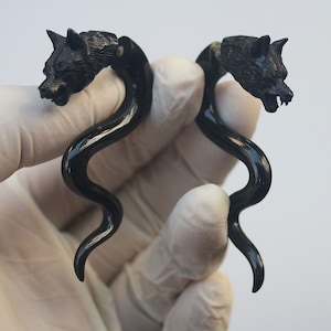 Op de afbeelding: Een paar zwarte oorstretchers in de vorm van een wolfskop. De wolfskoppen zijn gedetailleerd gesneden met vacht en open monden. De gauges hebben een gebogen, langwerpig ontwerp. De achtergrond is wit.