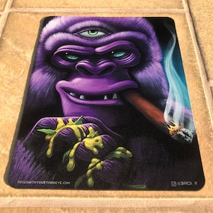 May include: Purple gorilla with a third eye, smoking a cigar and holding a green plant. The gorilla has a mischievous grin and is wearing a gold chain. The background is a dark blue with a white smoke plume. The text "focuswithyourthirdeye.com" is at the bottom left corner and "@3rd1_11" is at the bottom right corner.