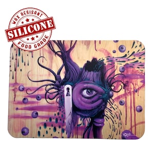 May include: A purple and blue abstract painting with a large eye in the center. The background is a light wood grain with purple and blue paint splatters. The painting is framed with a black border.