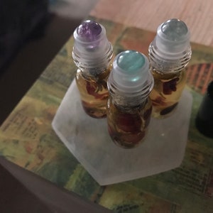 Puede incluir: Tres pequeños frascos de vidrio con rodillo llenos de un líquido dorado y elementos florales, coronados con tapas transparentes y rodillos de piedras preciosas en tonos morados y verdes. Los frascos están sobre una bandeja hexagonal blanca.