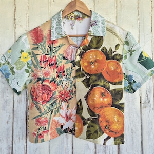 May include: A short-sleeved button-down shirt featuring a split-panel design. The left side displays vibrant red protea and Australian wildflowers on a tan background, while the right side features orange citrus fruit with green leaves on a cream base.