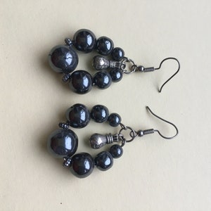 May include: A pair of dark gray beaded earrings. Each earring features a cluster of dark gray beads in varying sizes, connected by silver-toned metal components. The earrings have a hook closure.