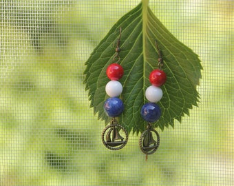 May include: A pair of dangle earrings with red, white, and blue beads and a sailboat charm. The earrings are hanging from a green leaf.