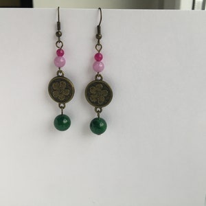 May include: A pair of dangle earrings with a green and pink bead design. The earrings feature a bronze flower charm and a green bead at the bottom.